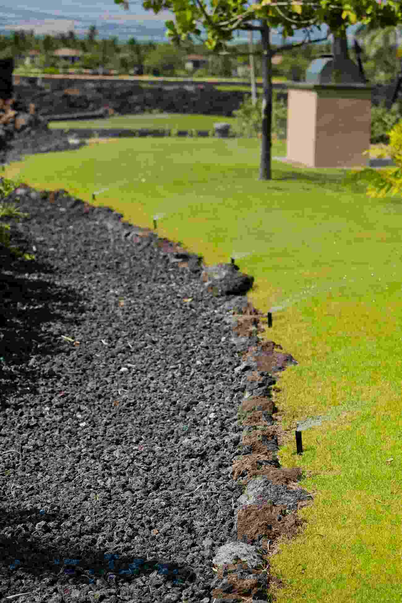 A section of a garden with a black gravel border and green grass, featuring sprinkler heads spraying water onto the lawn.