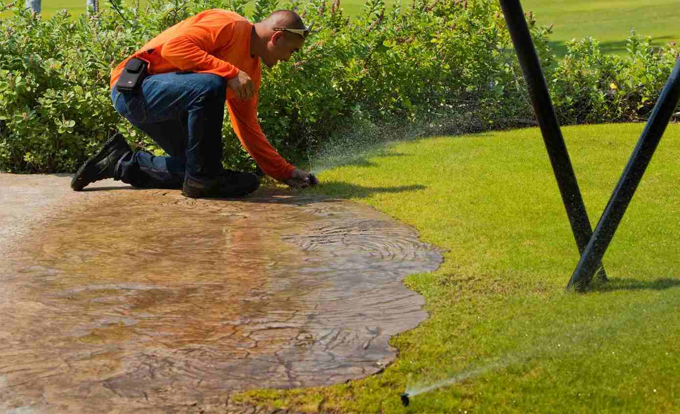 A man in an orange shirt kneeling on a lawn while adjusting a sprinkler system, with water spraying onto the grass.