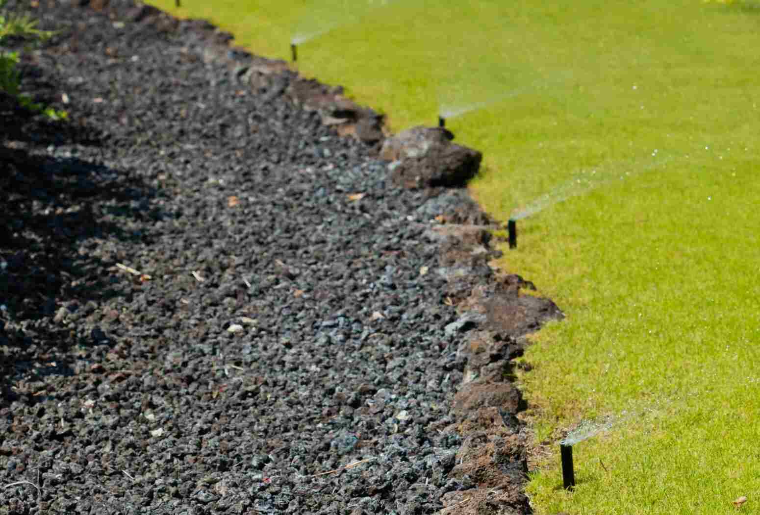 A section of a garden with a black gravel border and green grass, featuring sprinkler heads spraying water onto the lawn.