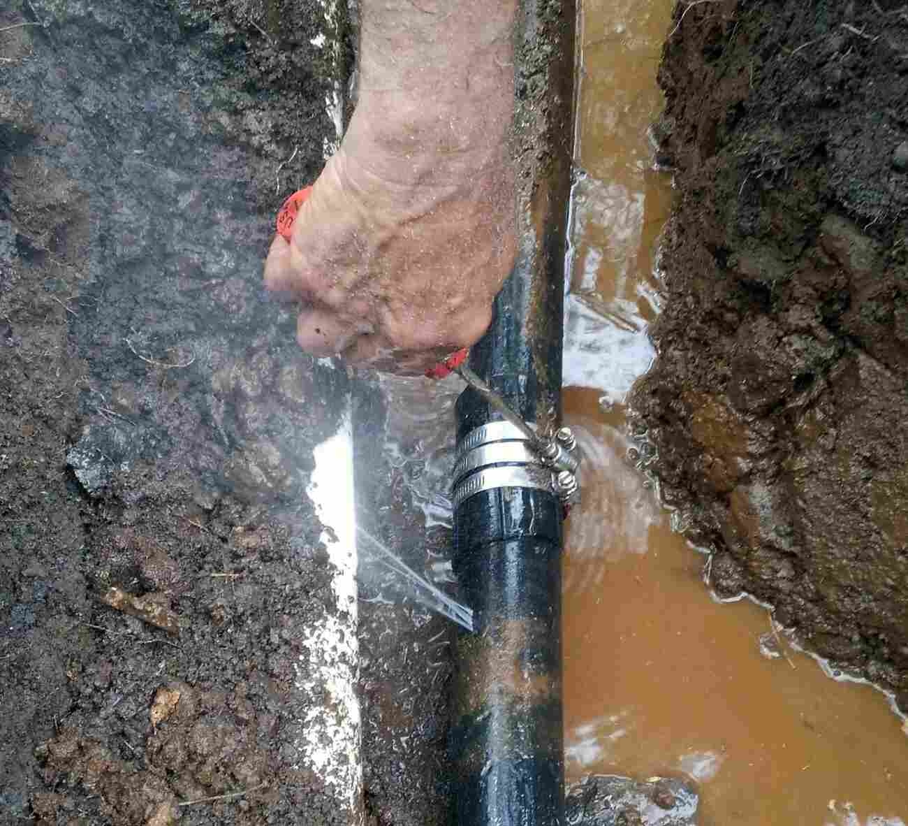 A person's hand is working on a pipe in a trench with water and mud around it.