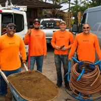 Four workers in bright orange shirts standing with a wheelbarrow and equipment, smiling at the camera. In the background, there are vehicles and coils of hose.