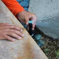 A person adjusting a sprinkler head on a lawn, with their hands visible on a stone surface.