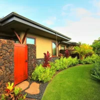 A side view of a house surrounded by lush greenery, with a stone wall and a wooden door visible.
